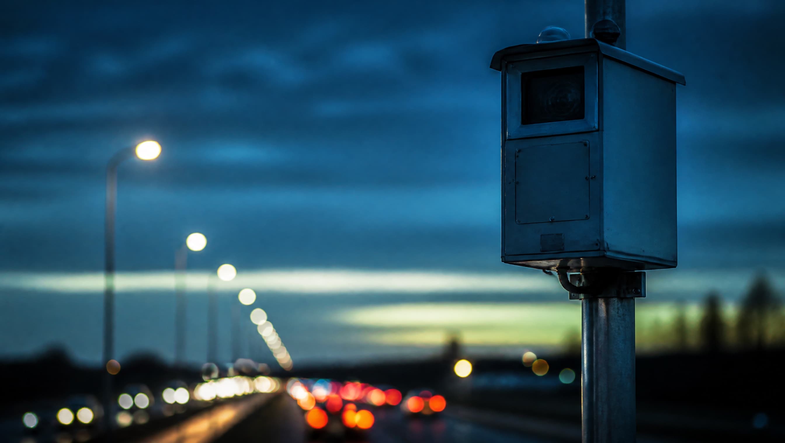 Illustrative roadside camera cabinet at dusk with traffic lights beyond